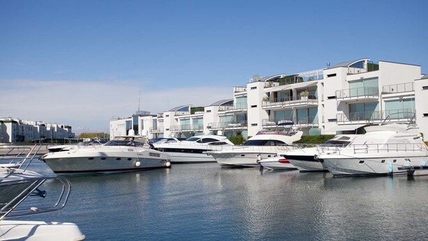 A marina with several yachts docked in front of modern buildings with balconies and large windows. - Libeccio (Jesolo)