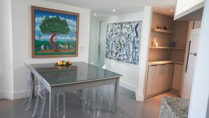 A dining area with a glass table and chairs, a painting of a tree, and a tiled backsplash. - Cobertura Luxor Paulo Miranda (João Pessoa)