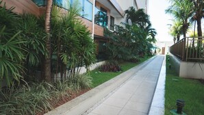 A modern building with a clean, tiled pathway flanked by lush greenery and palm trees. - Cobertura Luxor Paulo Miranda (João Pessoa)