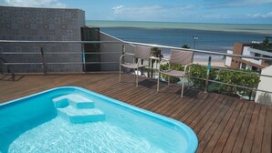 A pool area with a view of the ocean, wooden deck, and outdoor seating. - Cobertura Luxor Paulo Miranda (João Pessoa)