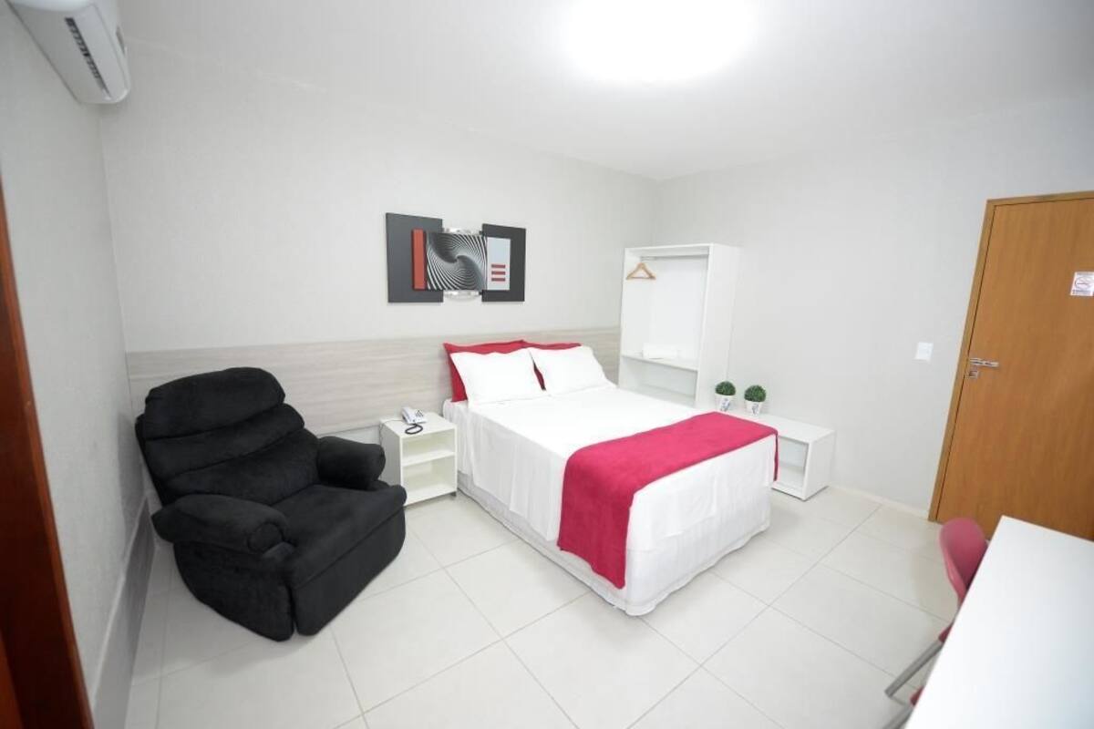 Una habitación de hotel con una cama, un sillón negro, una mesita de noche con un teléfono, un armario blanco y una obra de arte colgada en la pared.