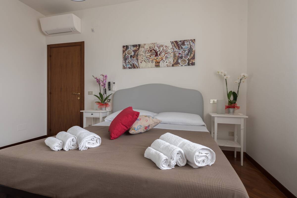 A neatly made bed with rolled white towels, a red pillow, and a floral-patterned pillow. There is a wall-mounted artwork above the bed and a small white nightstand with a potted plant beside it.