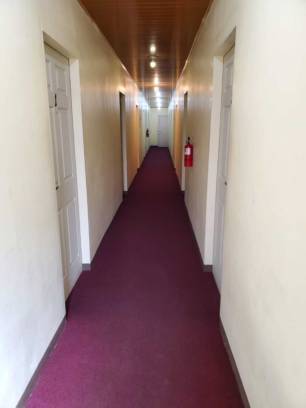 A long, narrow hallway with white walls and a red carpet. There are doors on both sides, and a fire extinguisher is mounted on the wall.