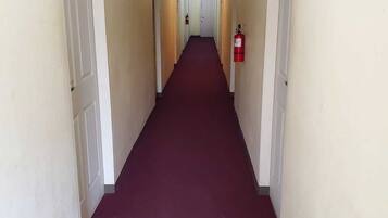 A long, narrow hallway with white walls and a red carpet. There are doors on both sides, and a fire extinguisher is mounted on the wall.