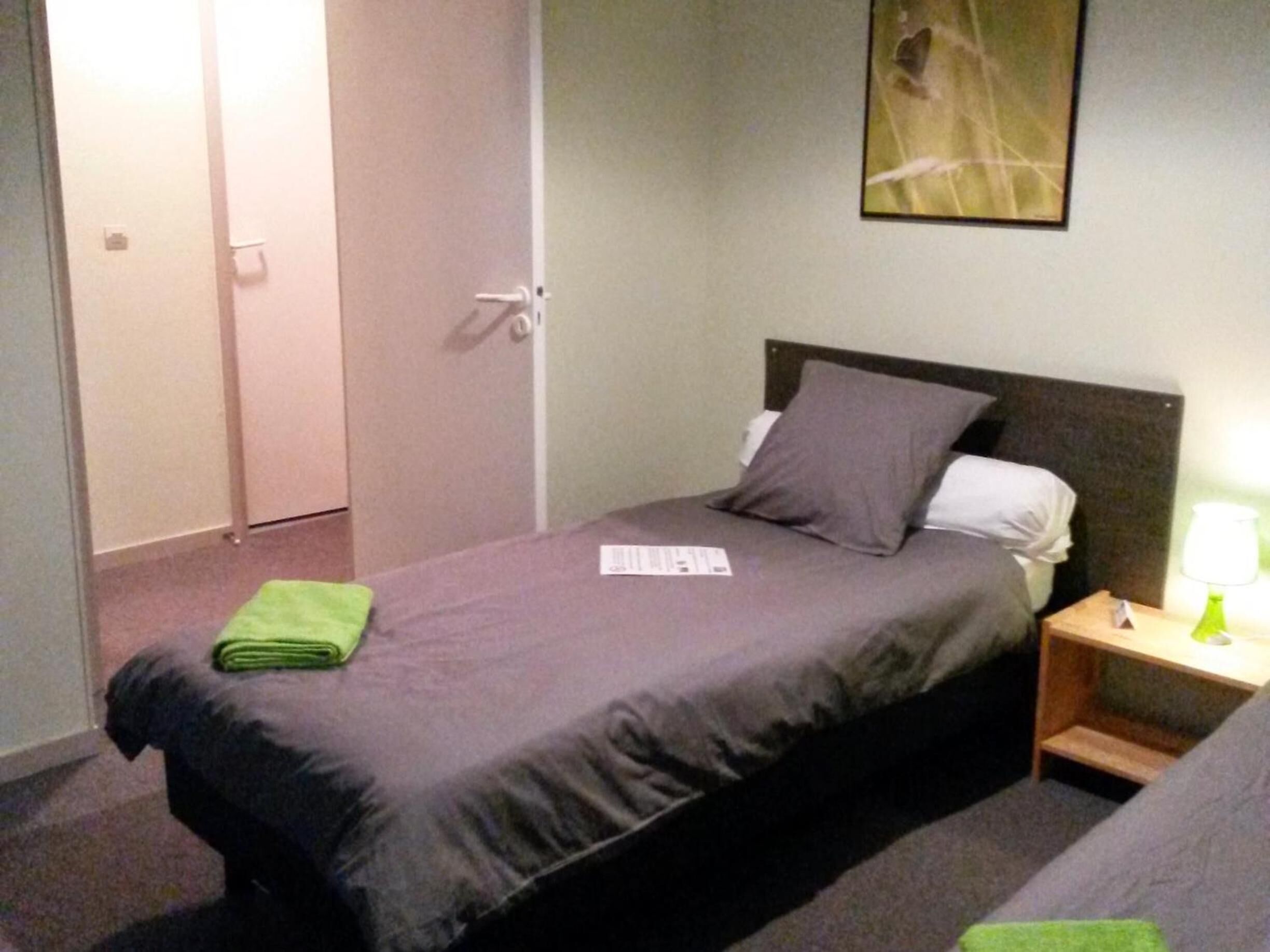 Un lit simple avec une couvre-lit et des oreillers gris, une serviette verte sur le lit, une table de chevet avec une lampe, et une porte sur la gauche.