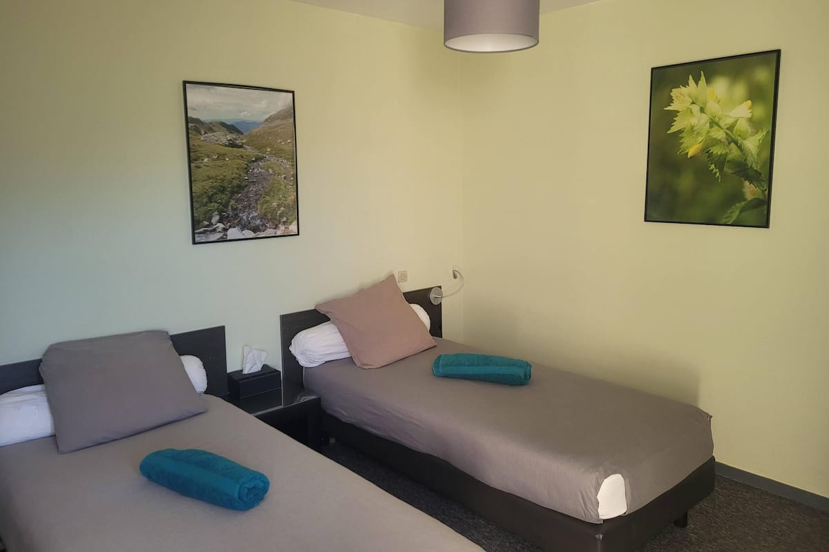 A hotel room with two single beds, a small bedside table with a lamp, and two framed pictures on the wall.