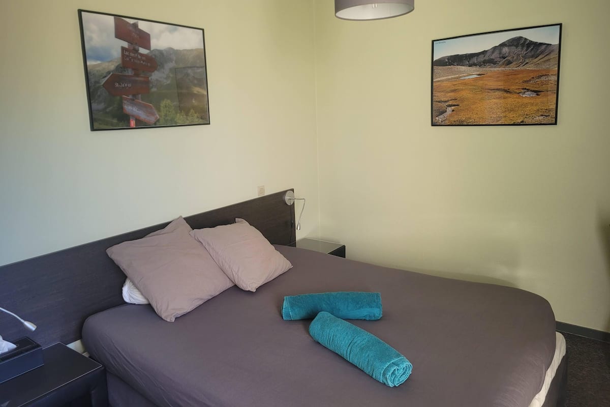 A neatly made bed with two pillows and two rolled towels, a bedside table with a lamp, and two framed pictures on the wall.
