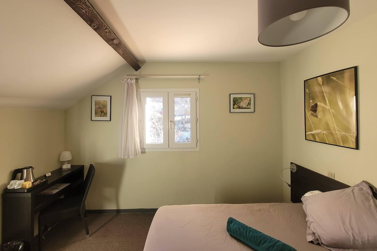 A bedroom with a bed, a desk, a chair, a window with curtains, and two framed pictures on the wall.