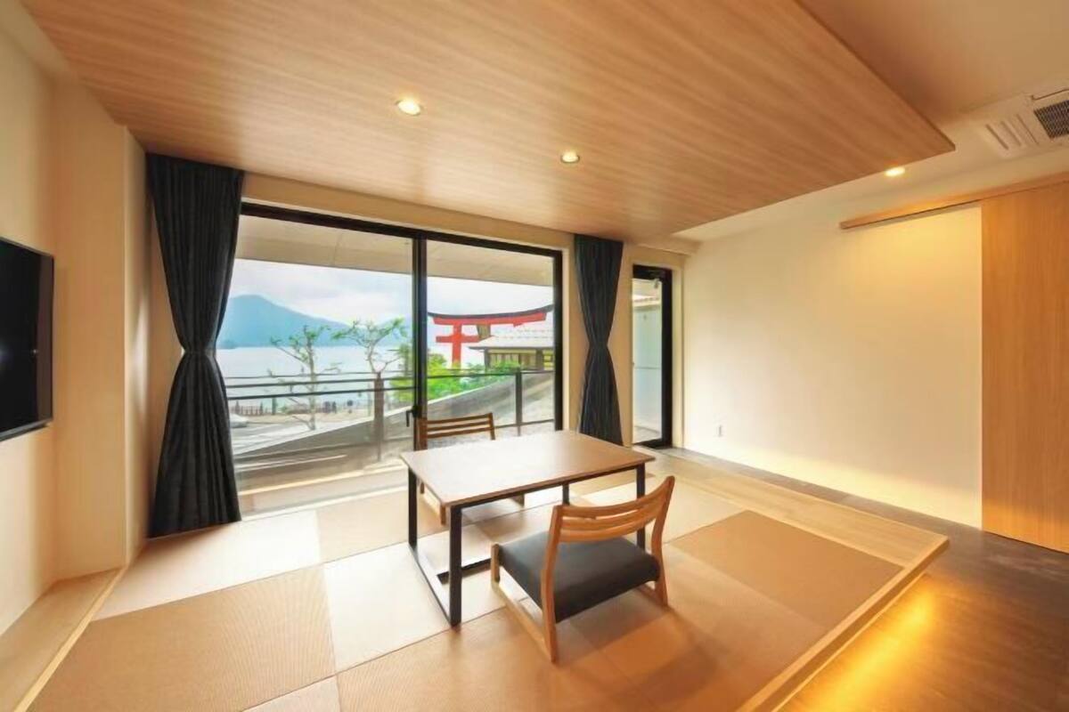 A modern hotel room with a wooden ceiling, a large window with curtains, a dining table, and a TV.
