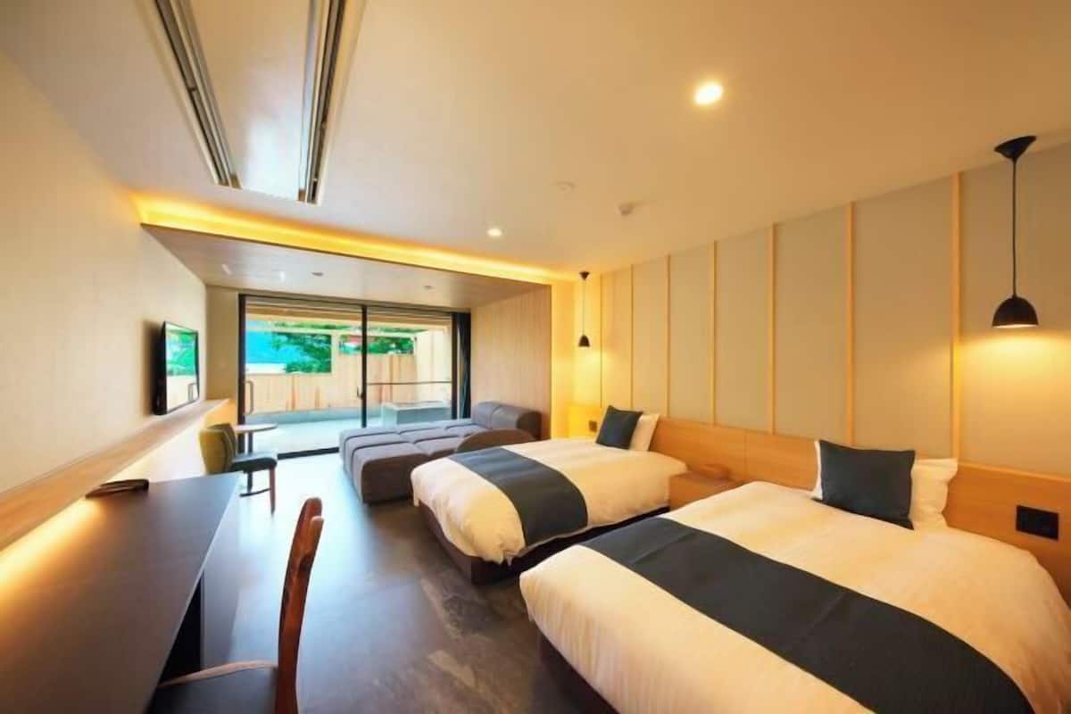 A modern hotel room with two beds, a dining area, and a view of the outdoors.