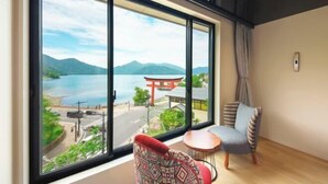 A room with a large window offering a view of a lake, mountains, and a traditional Japanese torii gate. There is a small table and a chair with patterned cushions. - Nikko Chuzenji-ko Onsen Hotel Hana-An (Nikko)