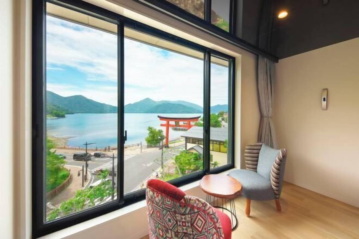 A room with a large window offering a view of a lake, mountains, and a traditional Japanese torii gate. There is a small table and a chair with patterned cushions.