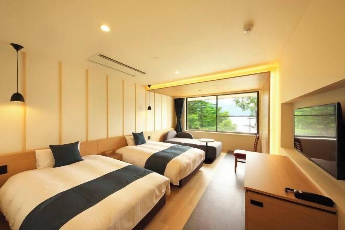 A hotel room with two beds, a desk, a chair, a TV, and a large window with a view of greenery.