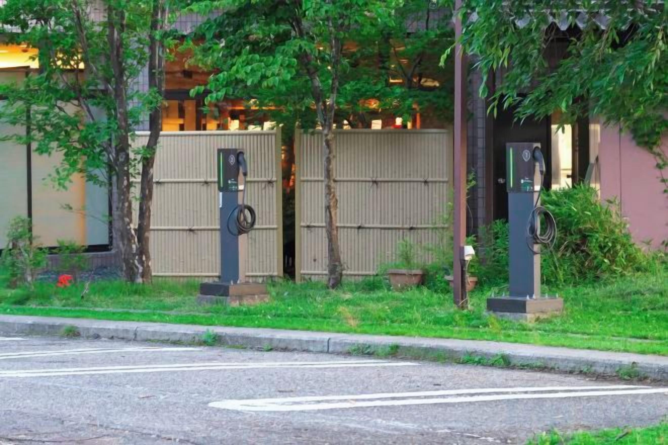 A parking area with electric vehicle charging stations, a paved road, and a building with a gated entrance in the background.
