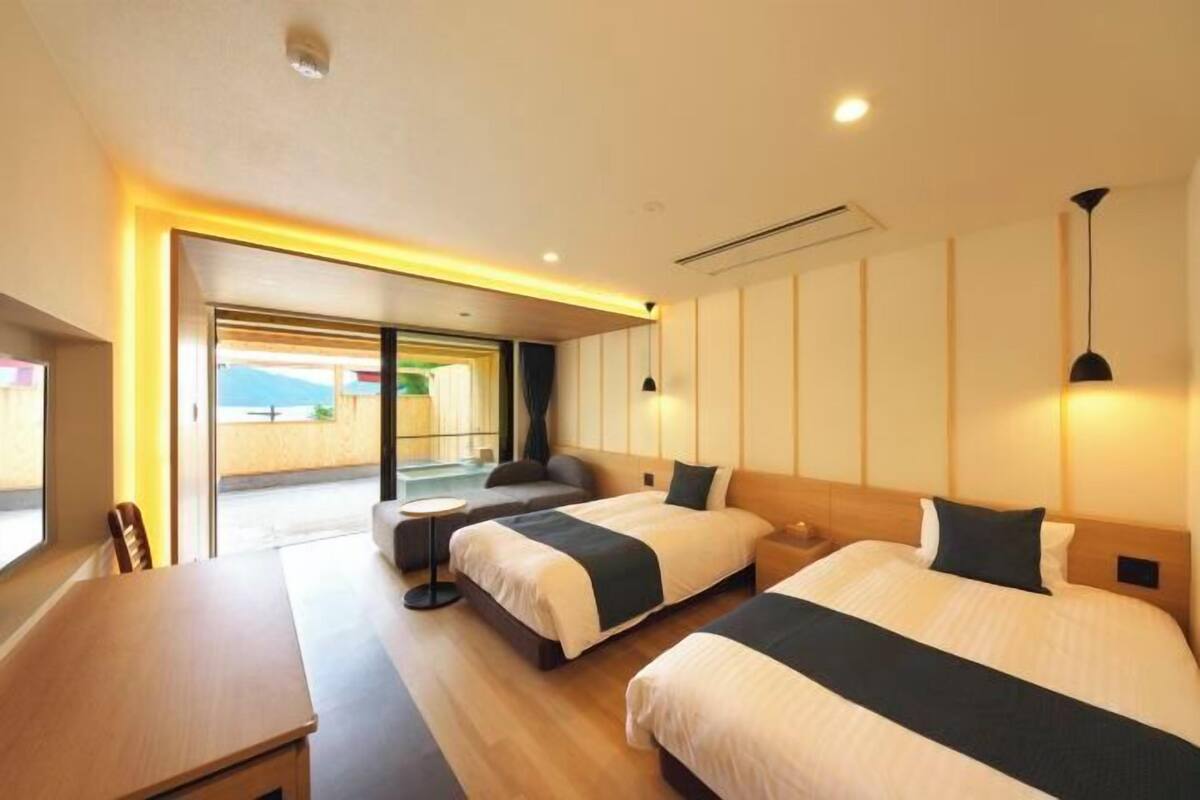 A hotel room with two beds, a small table, and a sofa. There is a large window providing a view of the outdoors.