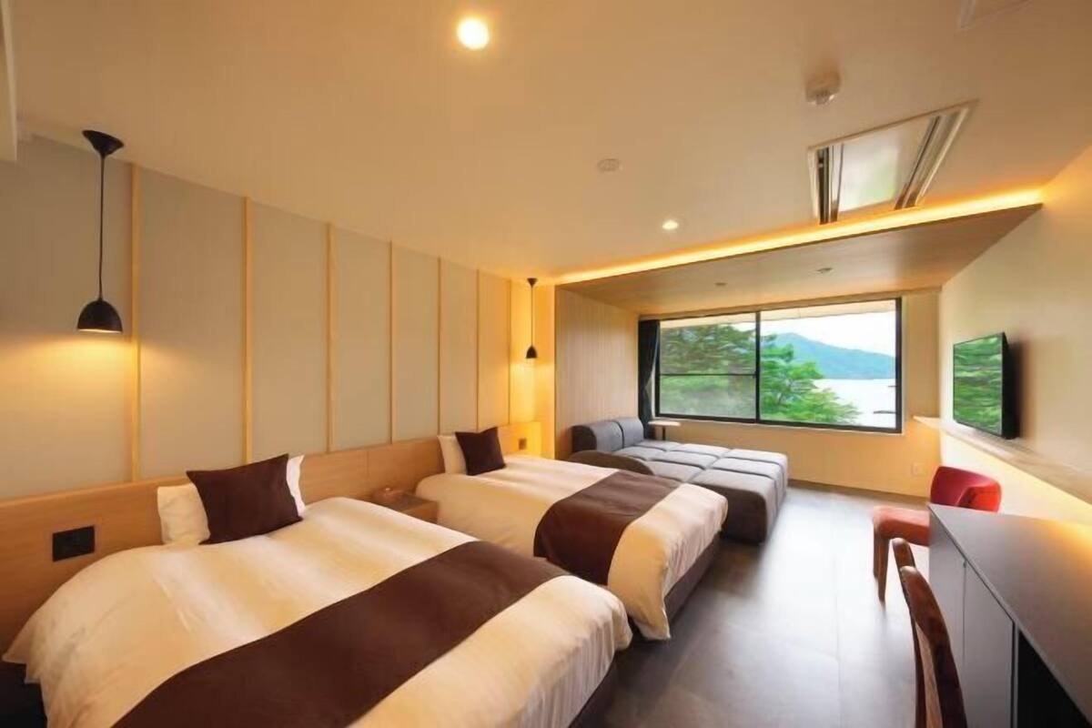 A modern hotel room with two beds, a seating area, a TV, and a large window with a view.