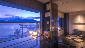 A modern hotel room with a large window overlooking a body of water and mountains, a glass partition, and a minimalist interior design.