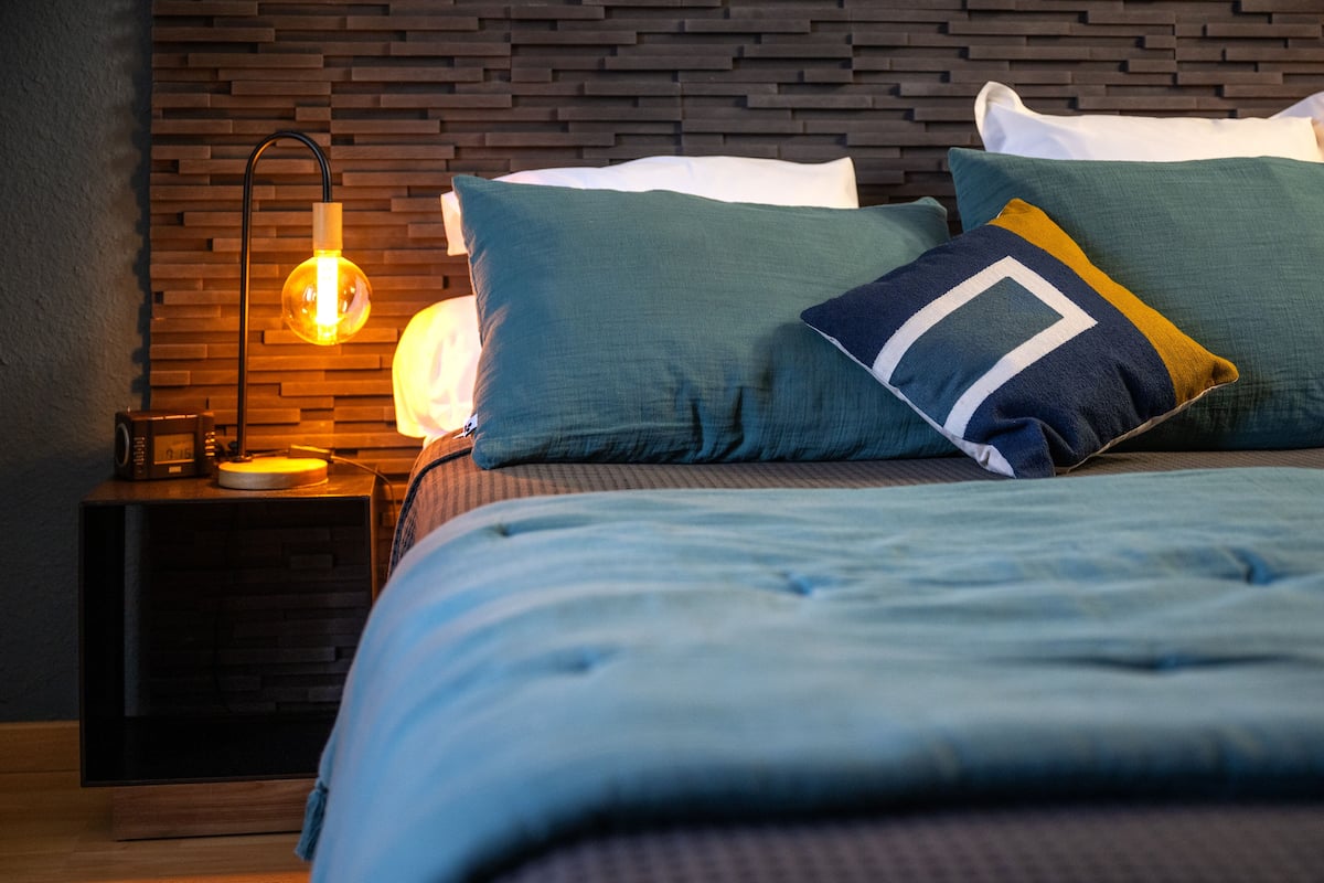 A neatly made bed with a blue comforter and pillows, a wall-mounted lamp, and a dark wooden headboard.