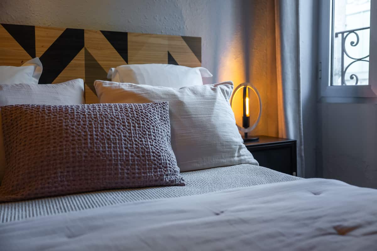 A neatly made bed with multiple pillows, a wooden headboard with geometric patterns, a bedside lamp, and a window with a metal railing.