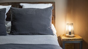 Una cama bien hecha con ropa de cama y almohadas grises, una mesita de noche de madera con una lámpara encendida y un cabecero de madera.