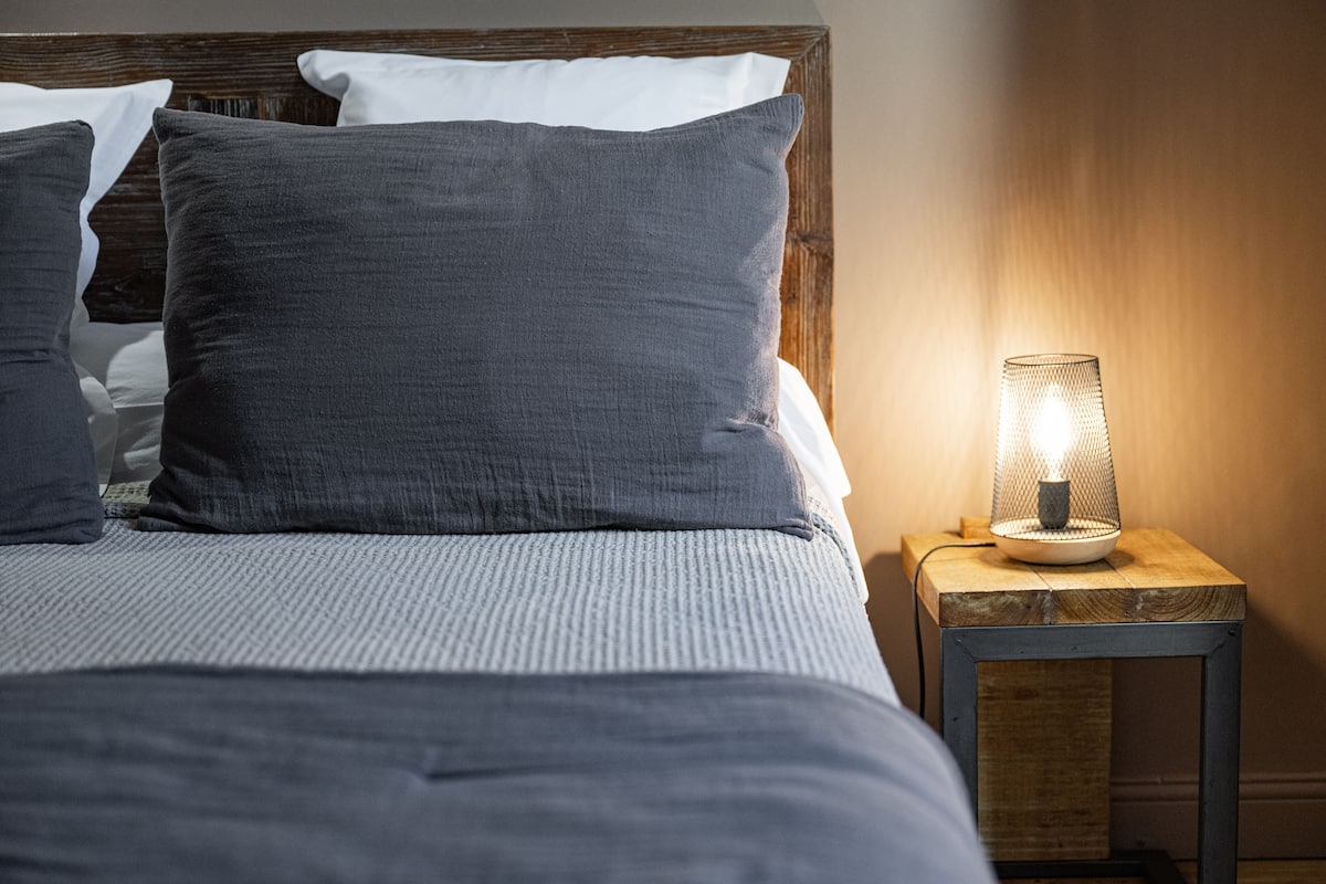 A neatly made bed with grey bedding and pillows, a small wooden nightstand with a lit lamp, and a wooden headboard.