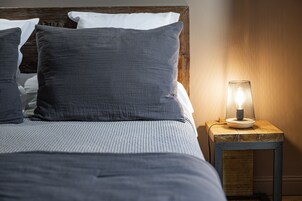 Una cama bien hecha con ropa de cama y almohadas grises, una mesita de noche de madera con una lámpara encendida y un cabecero de madera.