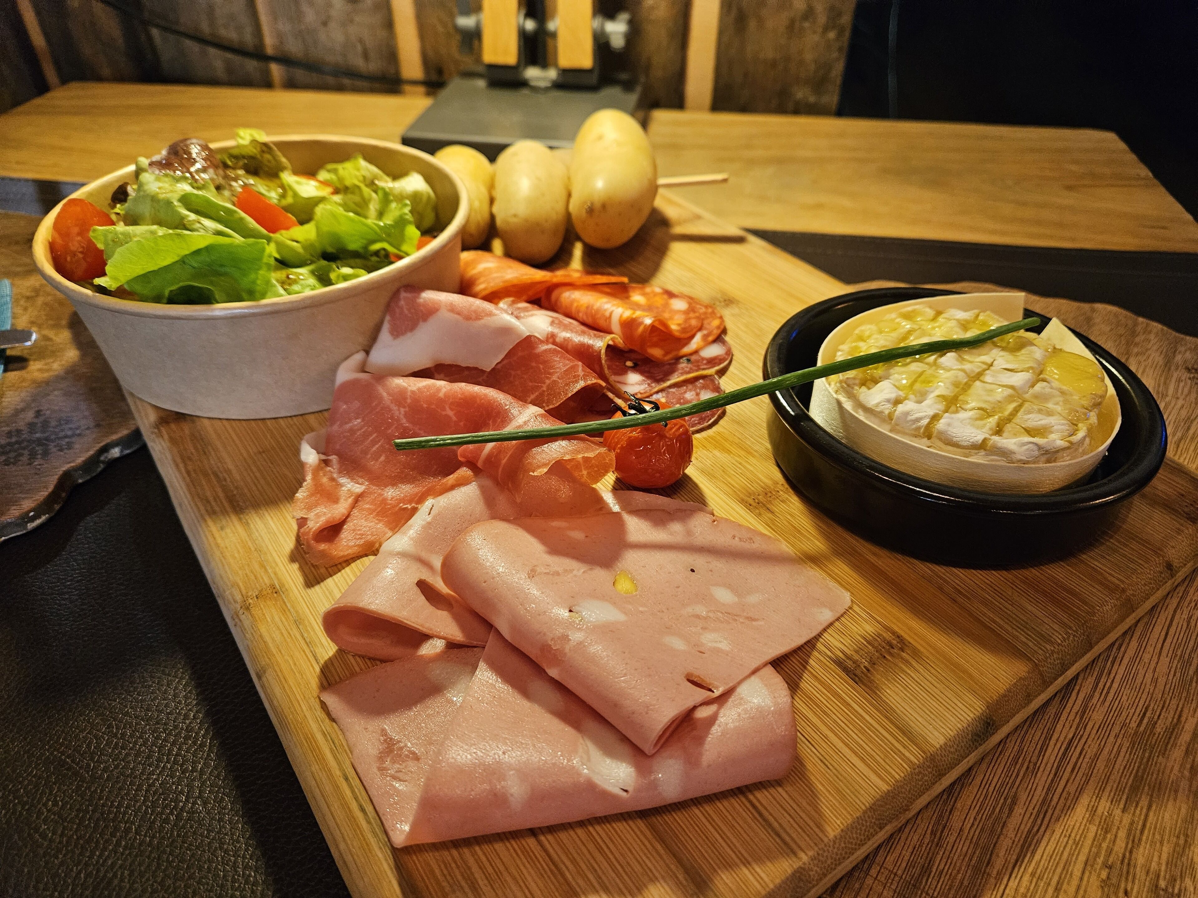A wooden board with various food items including sliced meats, a bowl of salad, and a dish of cheese.