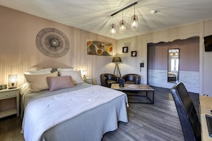 A modern hotel room with a large bed, a desk, a chair, and a sofa. There is a painting on the wall and a lamp on the desk.