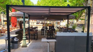 Un café al aire libre con mesas y sillas bajo un toldo sombreado, rodeado de follaje y una fuente de piedra.