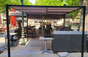 Un café al aire libre con mesas y sillas bajo un toldo sombreado, rodeado de follaje y una fuente de piedra.