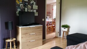 Une pièce avec une commode en bois, un téléviseur posé dessus, un petit tabouret, une plante en pot et un coin repas visible à travers une porte ouverte.