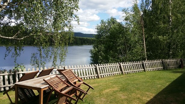 Une table et des chaises en bois, placées près d’un lac, avec une clôture en bois et des arbres en arrière-plan.
