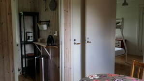 Une pièce avec une porte en bois, une petite étagère avec une bouilloire, une poubelle et un lit recouvert d’une courtepointe à motifs floraux.