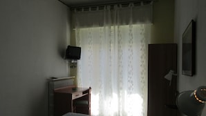 A room with a bed, a desk, a chair, a radiator, a television mounted on the wall, and a window with sheer curtains.