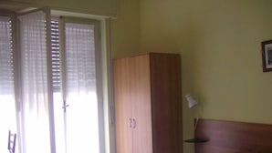 A bedroom with a bed, a wooden wardrobe, a desk with a chair, and a window with curtains.