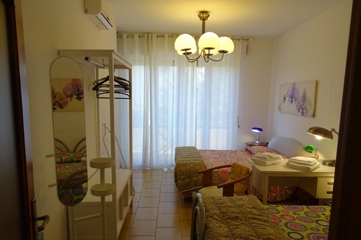 A hotel room with two beds, a desk with a lamp, a chair, a wardrobe, and a large window with curtains.