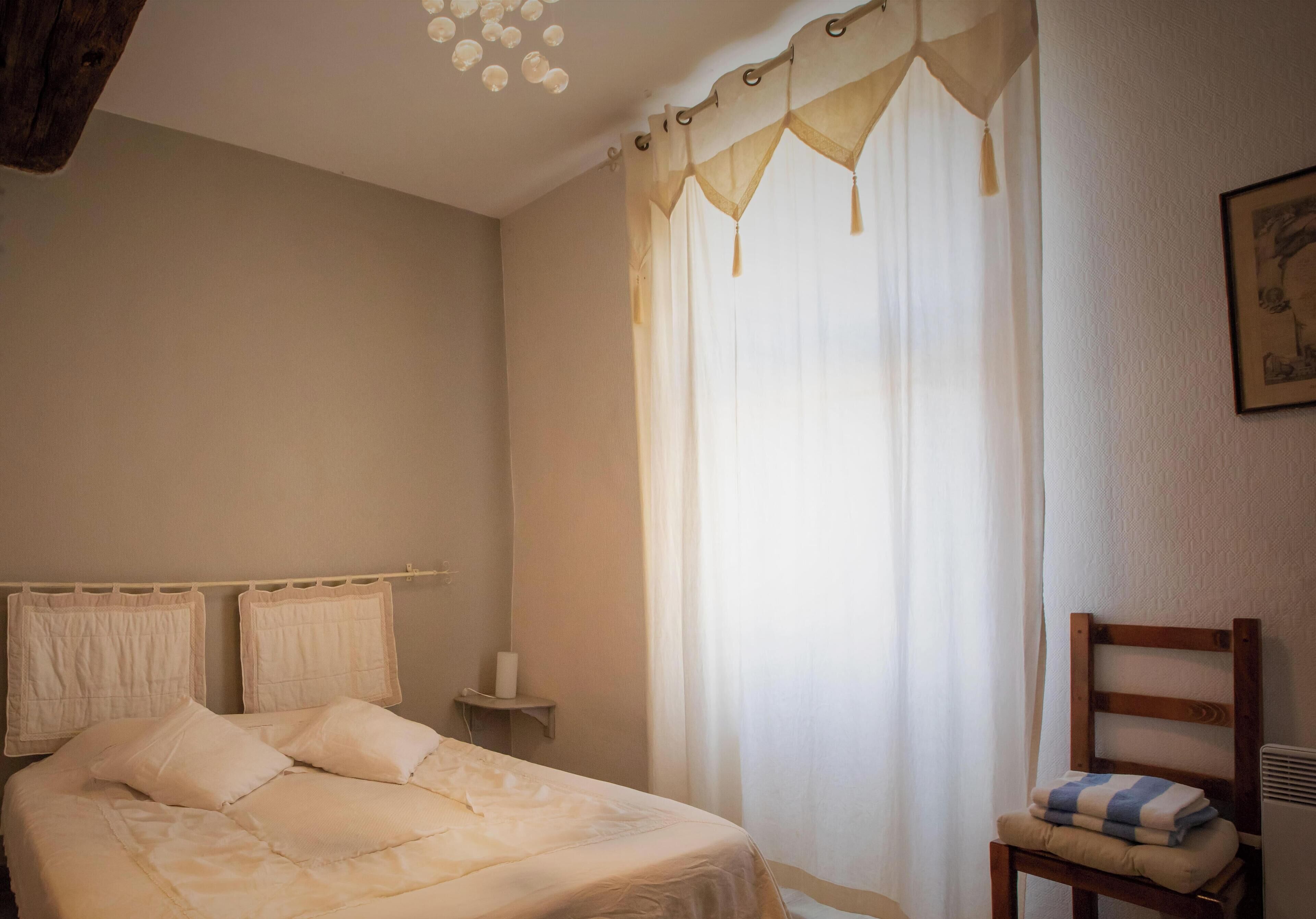 A bedroom with a bed, a chair, a bedside table, and a window with sheer curtains.