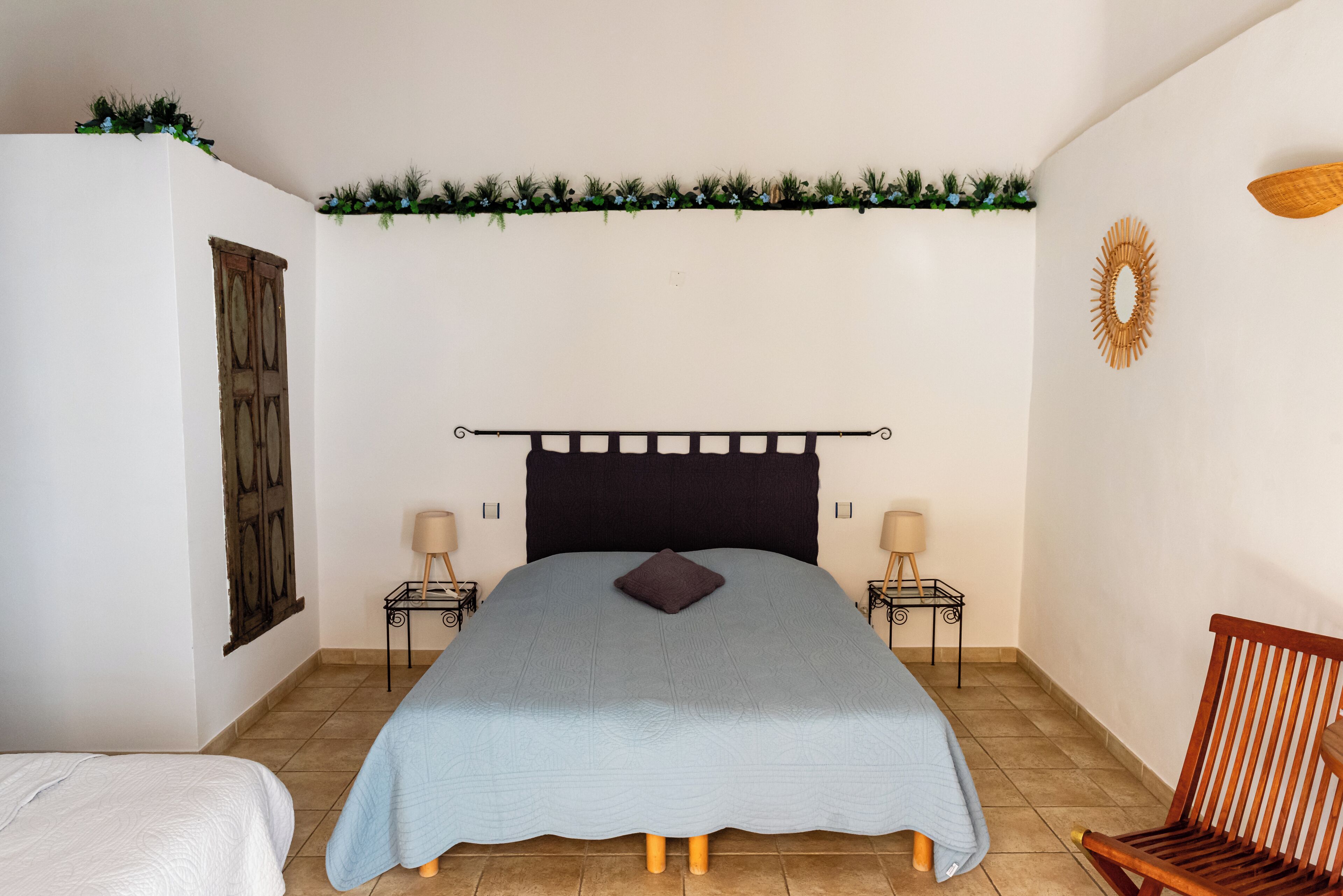 A bedroom with a bed, two bedside tables, a wooden chair, a decorative wall hanging, and a window with a wooden shutter.