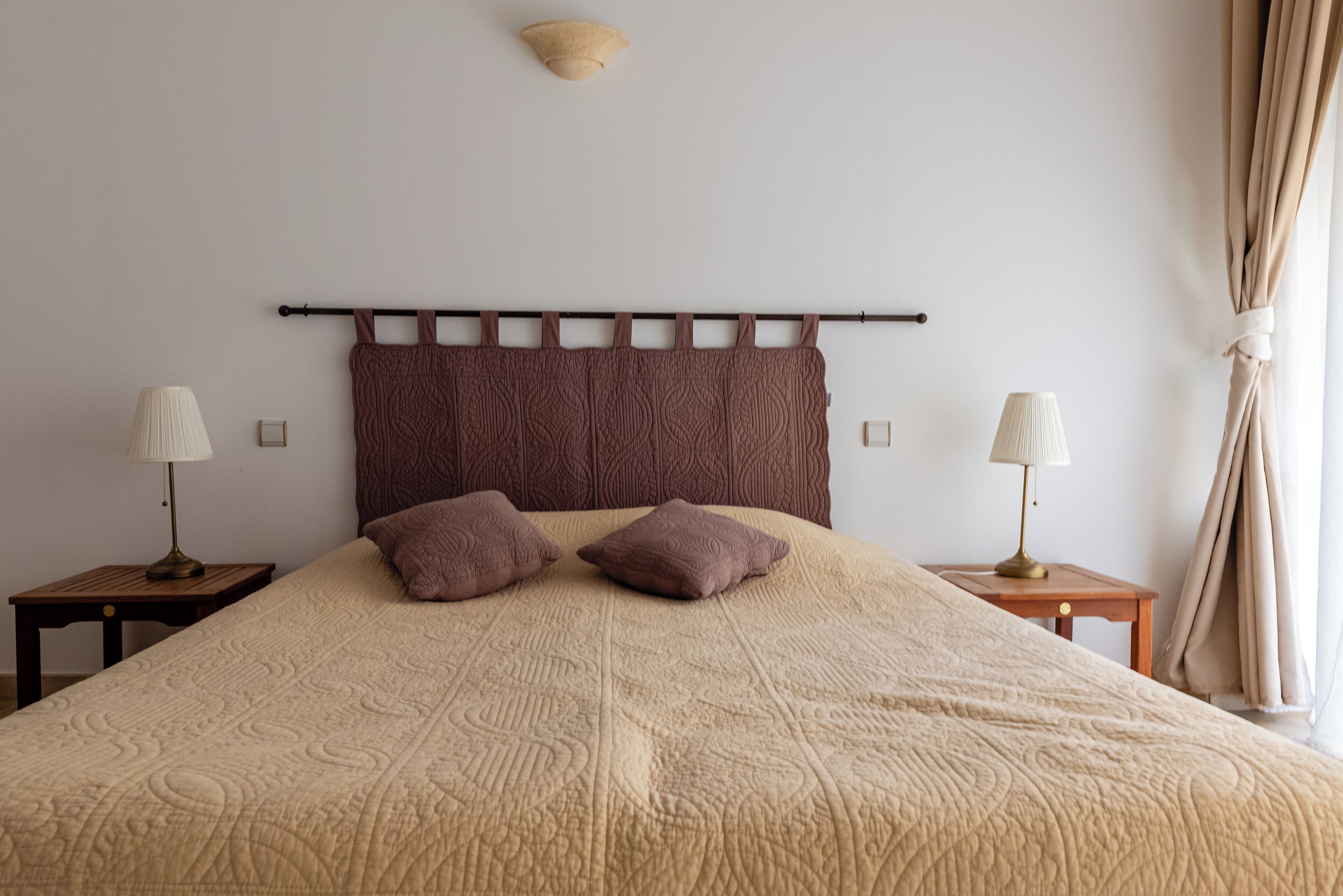 Un lit bien fait, recouvert d’une couverture beige à texture, avec deux oreillers assortis et une tête de lit ornée d’une barre horizontale. On y trouve deux tables de chevet, chacune agrémentée d’une lampe et d’un petit cadre photo.