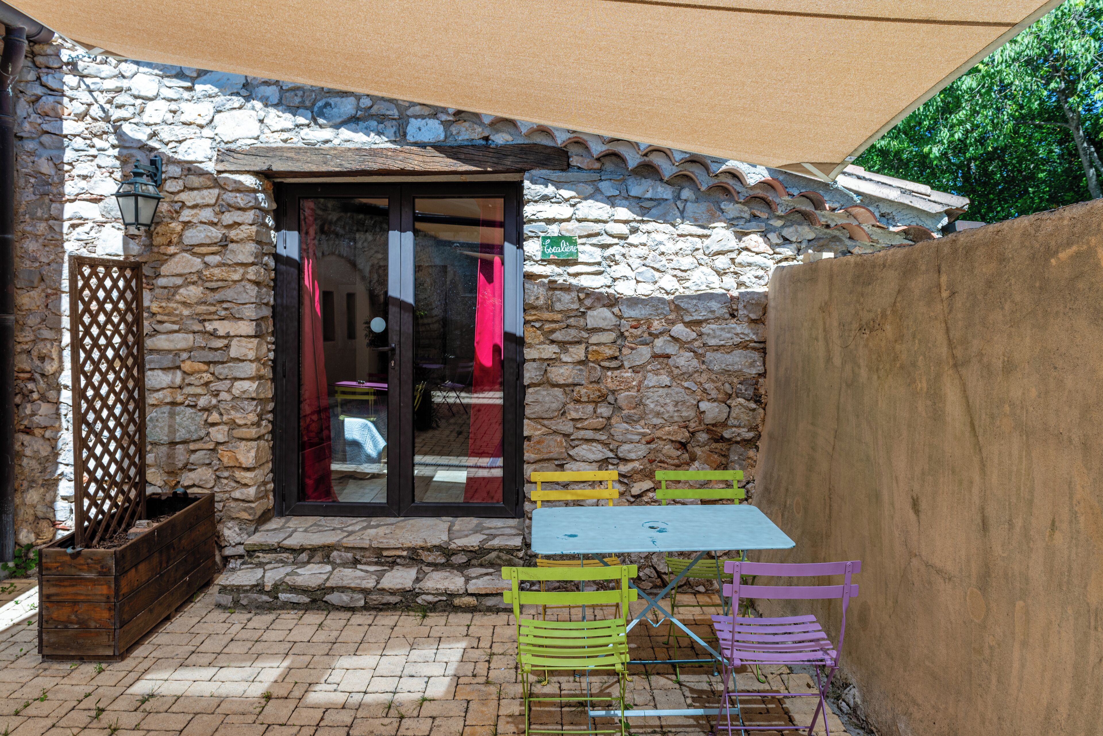 A stone building with a tiled roof, a wooden bench, and a table with colorful chairs.
