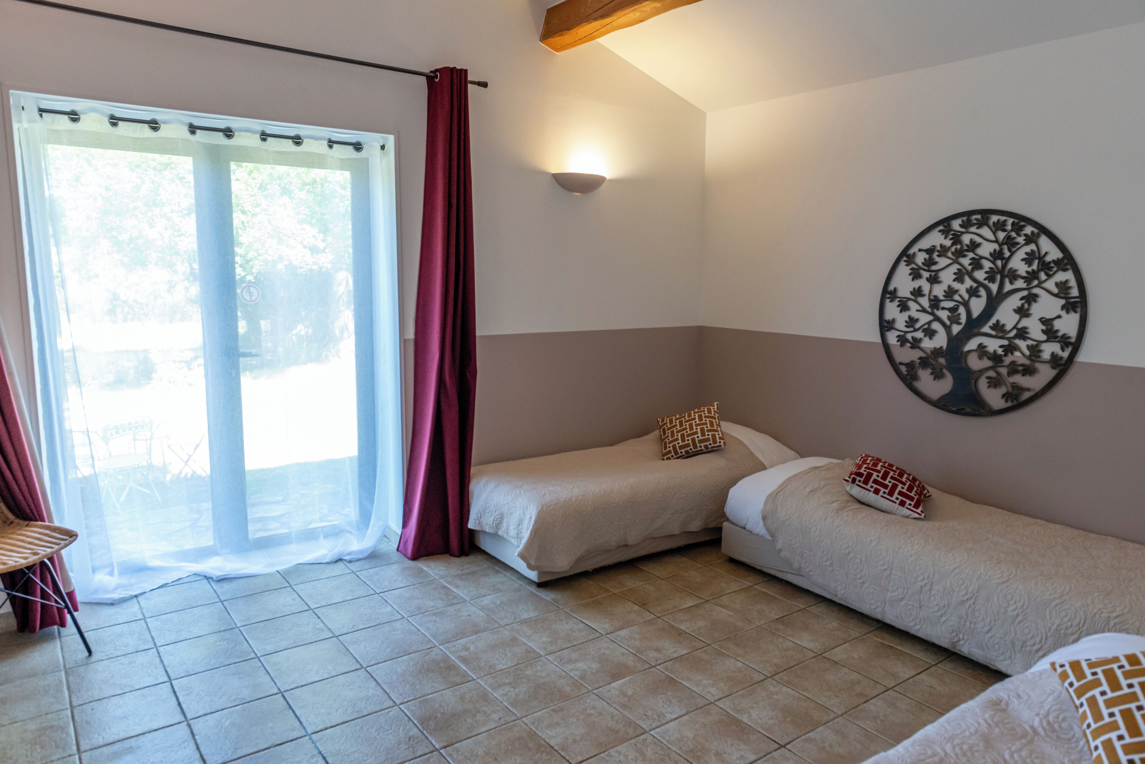 Een kamer met twee enkele bedden, een stoel, een tafel, een raam met gordijnen en een decoratie van een boom aan de muur.