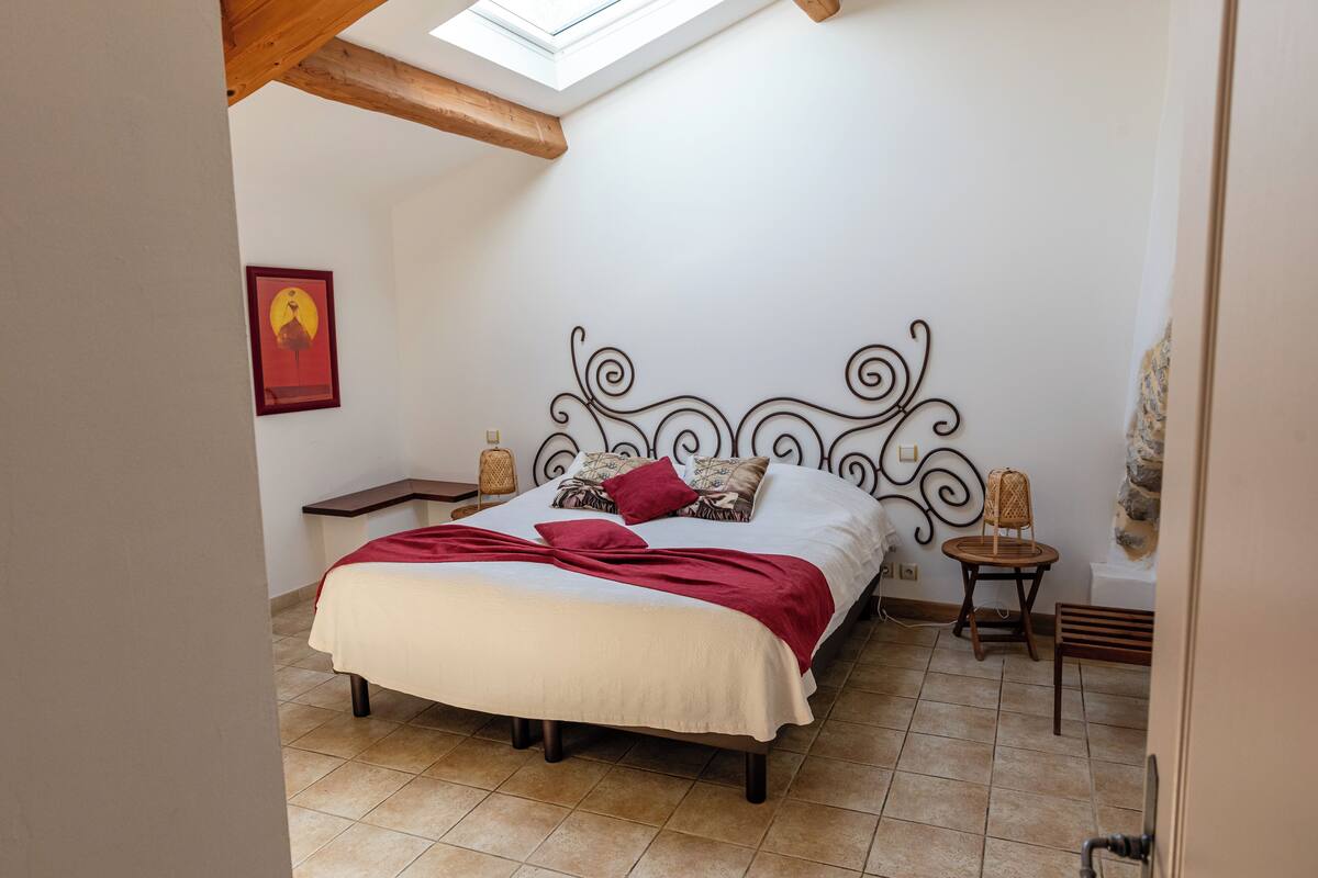 A bedroom with a bed, a skylight, a wall-mounted artwork, and a small bedside table with a lamp.