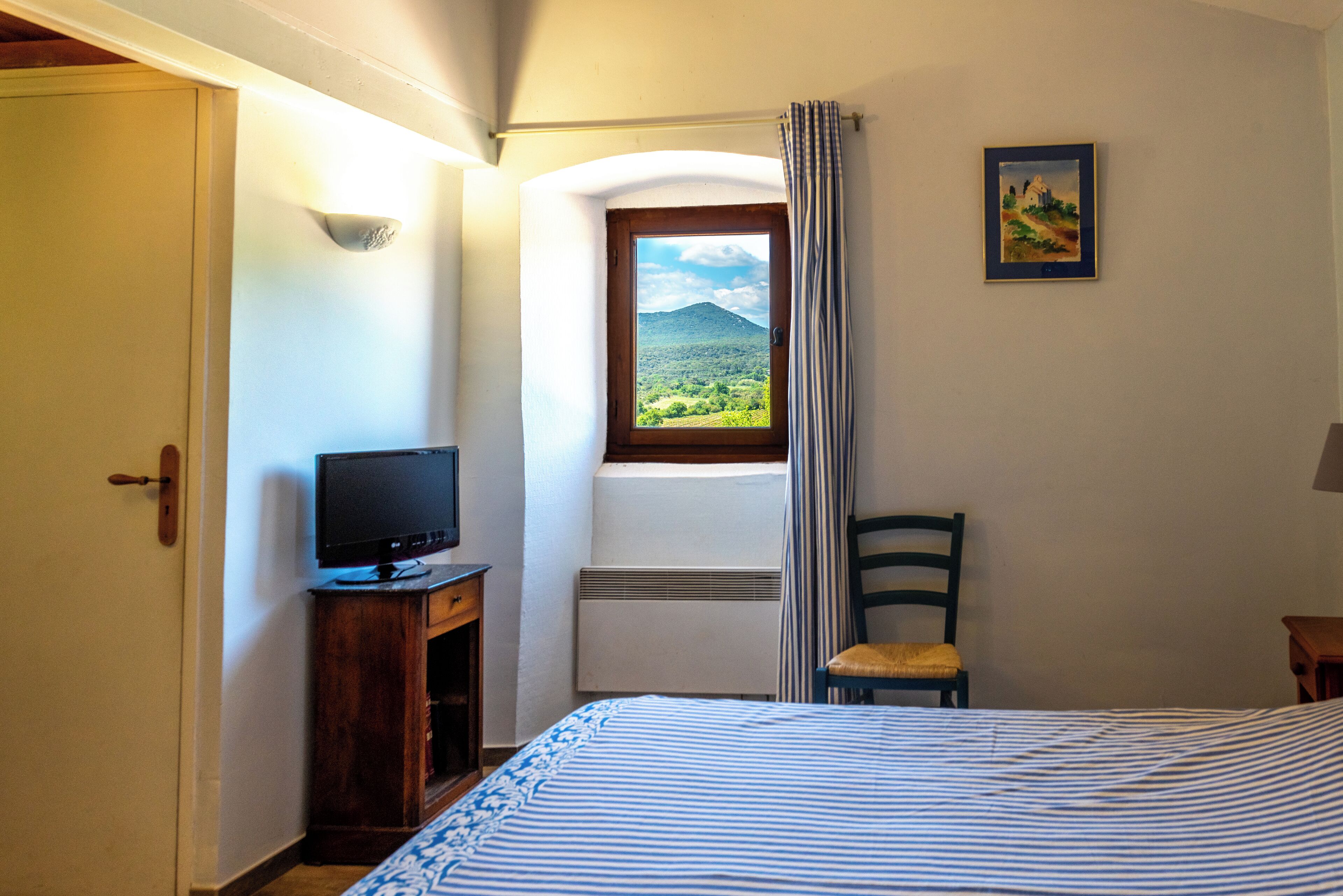 Una camera da letto con un letto, una televisione su un supporto in legno, una sedia, una finestra con vista su vegetazione e una montagna, e un quadro appeso al muro.