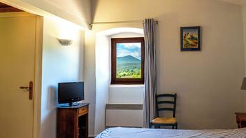 Una camera da letto con un letto, una televisione su un supporto in legno, una sedia, una finestra con vista su vegetazione e una montagna, e un quadro appeso al muro.