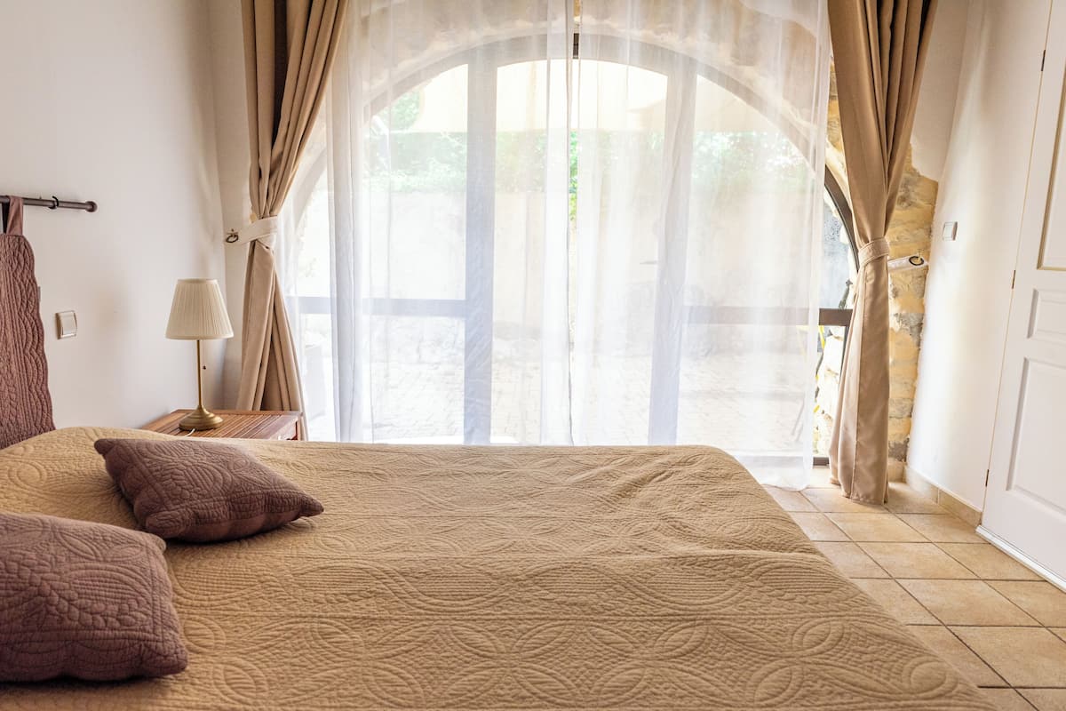 A bedroom with a large bed, beige curtains, a nightstand with a lamp, and a view through a glass door.