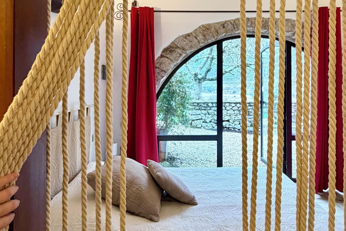A bed with a white bedspread and two pillows, a stone wall, and a view of trees through a window.