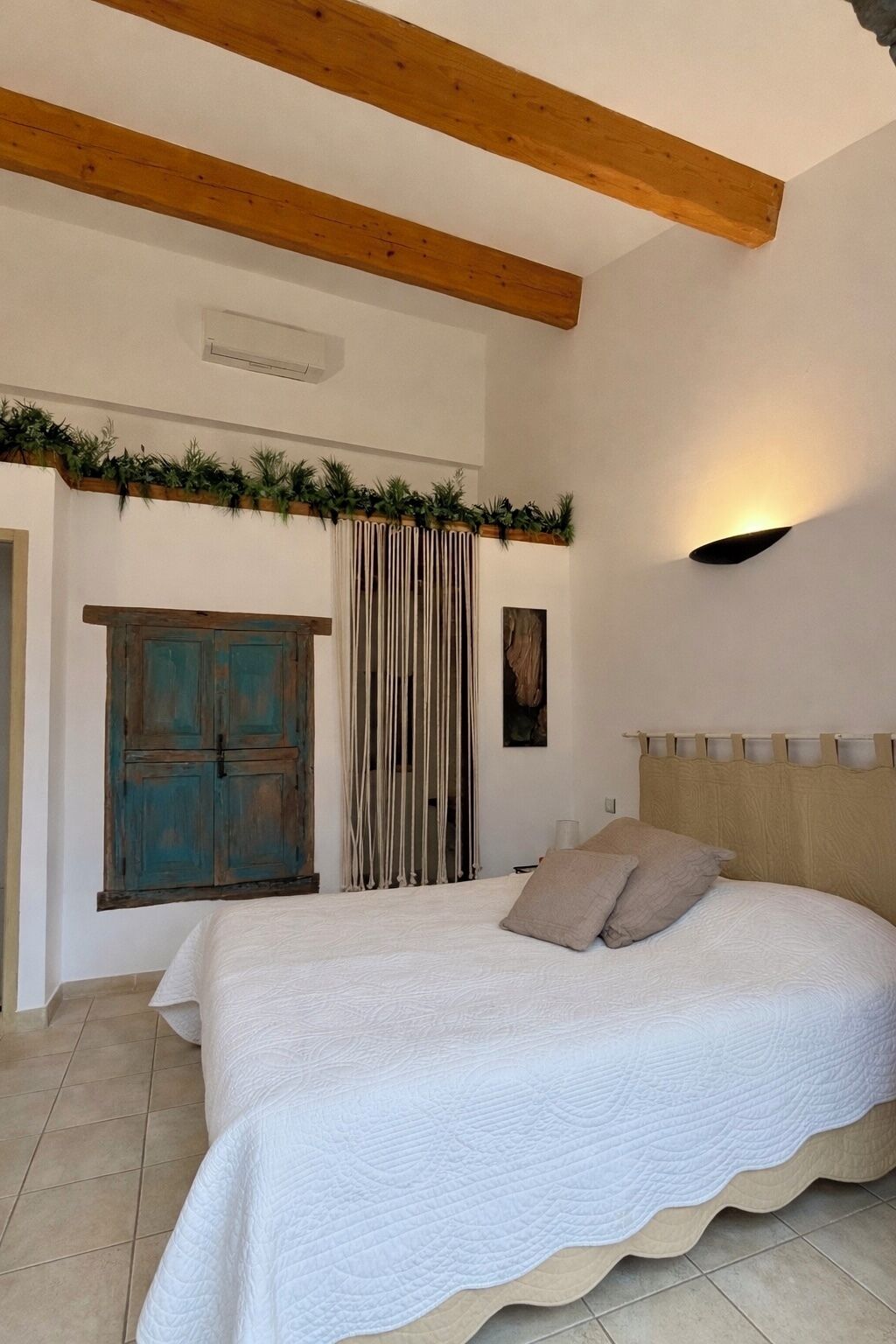 Une chambre à coucher avec un lit, des poutres en bois, un rideau, une lampe fixée au mur et une porte.