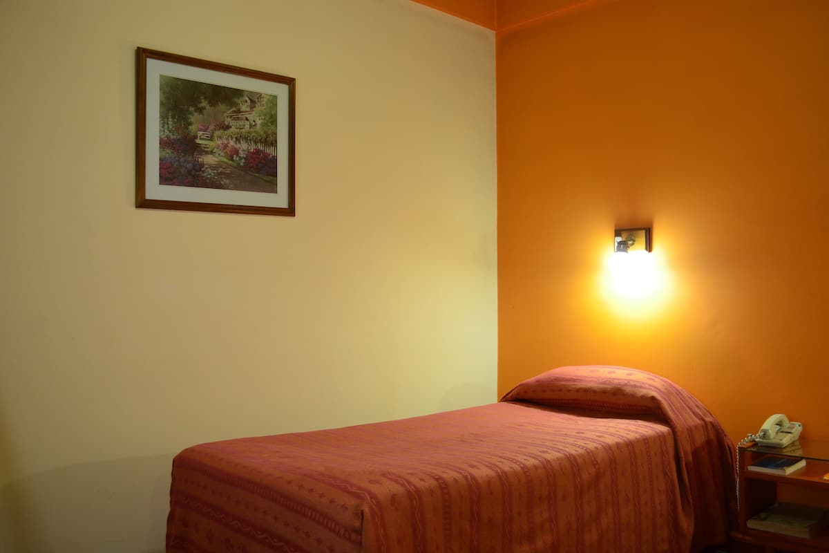 Un lit simple avec une couvre-lit rouge, dans une pièce où un tableau encadré est accroché au mur et où se trouve une lampe de chevet.