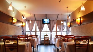 Un restaurante con mesas dispuestas para comer, una pantalla de televisión y grandes ventanales con cortinas.