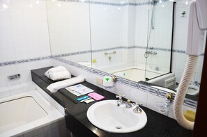 Casa de banho com banheira, lavatório e chuveiro. Há produtos de higiene no balcão, uma toalha e um espelho.